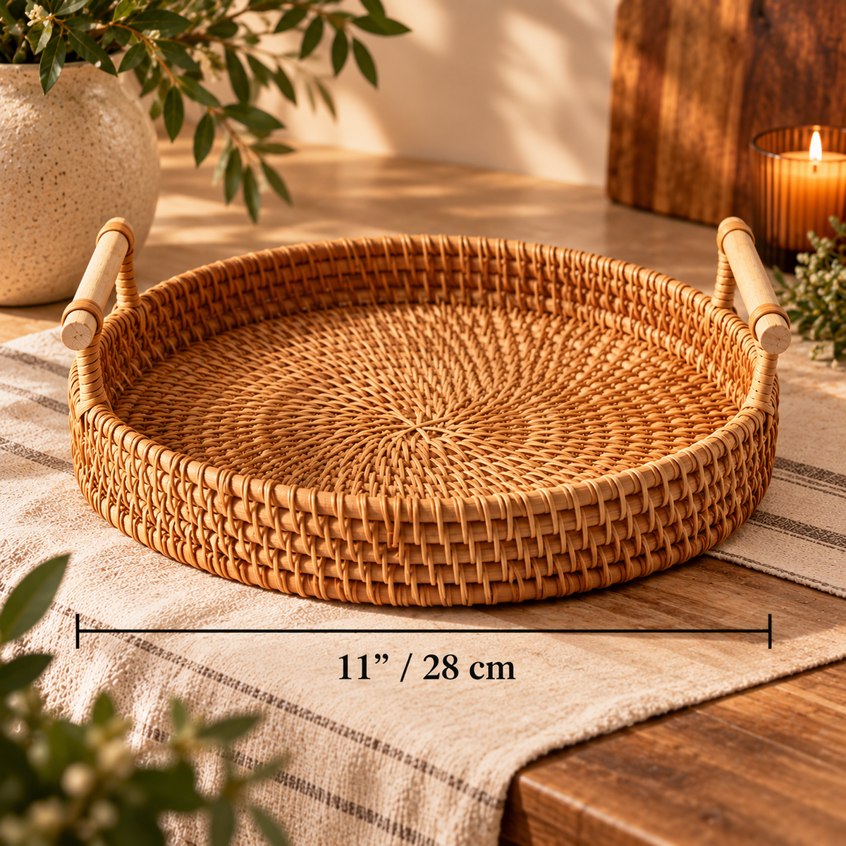 Round woven rattan tray with two handles, shown on a table runner with a candle and plants in a warm styled setting. A dimension overlay reads 11" / 28 cm across the tray.