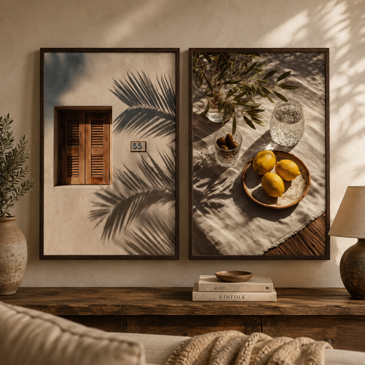 Two framed wall art prints hang above a wooden console table in a styled interior. The left print shows a beige wall with a brown shuttered window and palm leaf shadows, and the right print shows lemons on a tablecloth with glassware and leaf shadows.