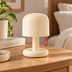 A small mushroom-shaped table lamp in a warm cream color sits on a wooden bedside table in a softly lit bedroom. A small red hangtag is attached to the side of the lamp.