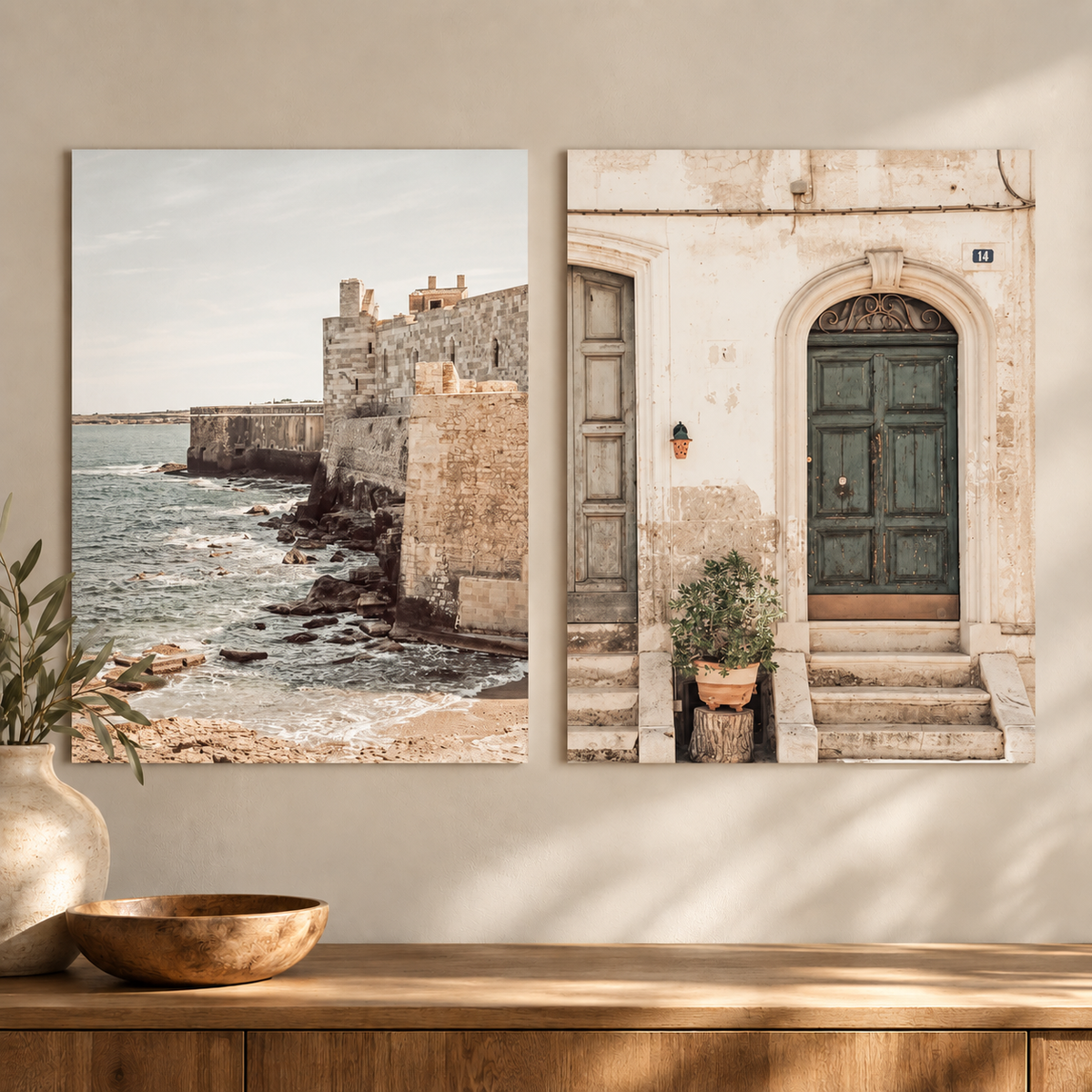 Two vertical wall art prints displayed side by side on a beige wall above a wooden console table. The left print shows a coastal stone fortress by the sea, and the right print shows a rustic building entrance with a green door and potted plant.