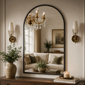 An arched black-framed wall mirror hanging above a wooden console table in a styled living room. The mirror reflects a chandelier and a sofa, while the room itself is decorated with two brass sconces, potted green plants in ceramic vases, and a lit candle on books.