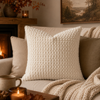 A square cream-colored textured throw pillow with a raised basketweave pattern, styled on a beige sofa in a cozy living room setting with a fireplace, candles, and a knit blanket.
