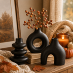 Three matte black ceramic vases with organic, sculptural shapes arranged on a wooden table in a cozy autumn setting. One vase holds a branch with orange berries.