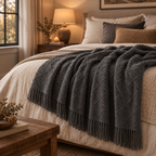 A dark charcoal gray knitted throw blanket with fringe trim draped over a bed in a warm, cozy bedroom setting.