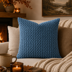 A textured blue throw pillow with a raised basketweave pattern, styled on a cozy beige sofa in a warm living room setting with a fireplace, candles, and a knit blanket.