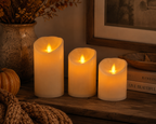Three faux flameless pillar candles in cream and off-white shades, arranged on a rustic wooden shelf with warm ambient lighting, dried flowers, a small pumpkin, and books in the background.