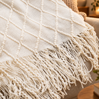 A close-up of a cream-colored woven throw blanket with a textured diamond pattern and long fringe, draped over a beige upholstered chair in a warm indoor setting.