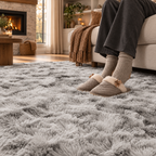 A person wearing gray lounge pants, beige socks, and taupe slip-on slippers sits on a light beige sofa in a cozy living room, with their feet resting on a large, shaggy gray area rug.