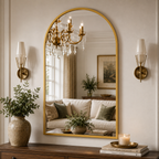 An arched gold-framed wall mirror with a crystal-accented reflection area, mounted on a cream wall in a styled living room setting flanked by two brass sconces and decorated with potted plants and books.