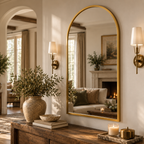 An arched gold-framed wall mirror hangs on a cream wall in a beautifully styled entryway, reflecting a cozy living room with a fireplace. The scene is decorated with a large ceramic vase of greenery, books, and brass sconces.