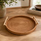 Round woven rattan tray with bamboo handles on a wooden table, styled with a plant and books in the background.