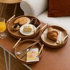 Three round woven rattan serving trays with light wood handles are arranged on a brown leather table in a cozy living room setting. The trays hold assorted breakfast items including a croissant, cookies, a slice of cake, a glass of orange juice, and a card.