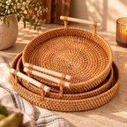 Three nested round woven rattan trays with light wood handles, styled on a stone surface with a vase of flowers and a candle in the background.