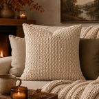 A square cream-colored textured throw pillow with a basketweave pattern, styled on a cozy beige sofa in a warm living room setting with candles, a knit blanket, and a fireplace in the background.