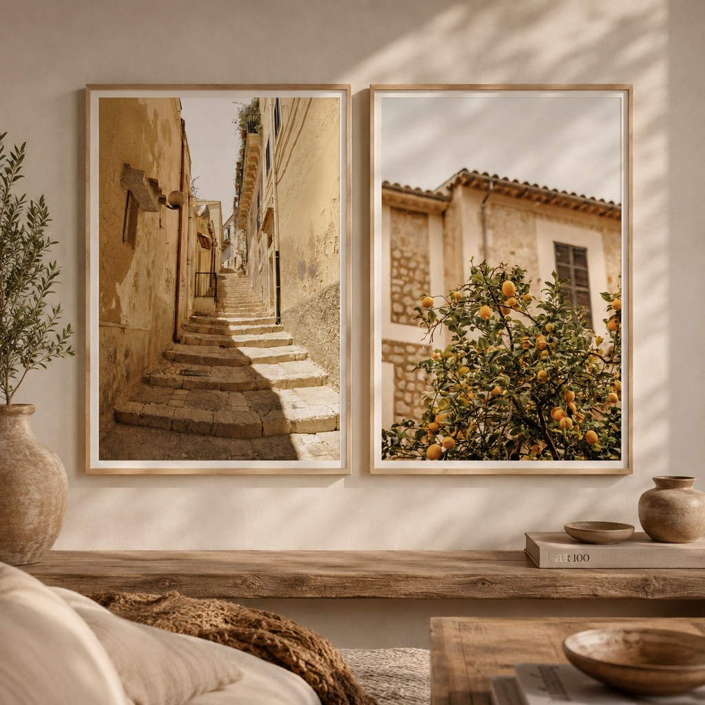 Two framed wall art prints hang on a beige wall above a wooden console table. The left print shows a sunlit stone staircase between warm-toned buildings, and the right print shows an orange tree in front of rustic buildings.