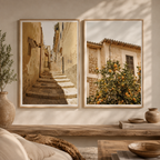 Two framed wall art prints hang on a beige wall above a wooden console table. The left print shows a sunlit stone staircase between warm-toned buildings, and the right print shows an orange tree in front of rustic buildings.