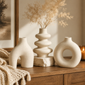 Three matte ceramic vases in a warm neutral beige color, styled on a wooden console table with dried pampas grass in the center vase, a cream knit throw blanket, and a lit candle in the background.