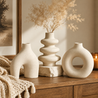 Three matte ceramic vases in a warm neutral beige color, styled on a wooden console table with dried pampas grass in the center vase, a cream knit throw blanket, and a lit candle in the background.