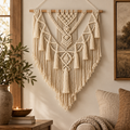 A large cream macrame wall hanging with intricate woven patterns and long tassels, suspended from a wooden rod in a warm, styled room setting with a window, plant, and sofa visible.