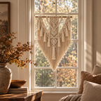 A cream macrame wall hanging with intricate woven patterns and tassels, suspended from a wooden rod in a sunlit room by a window with outdoor foliage visible behind it.