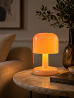 A glowing mushroom-shaped table lamp with a warm orange light, placed on a round marble side table in a cozy living room setting next to a sofa and a dark glass vase.
