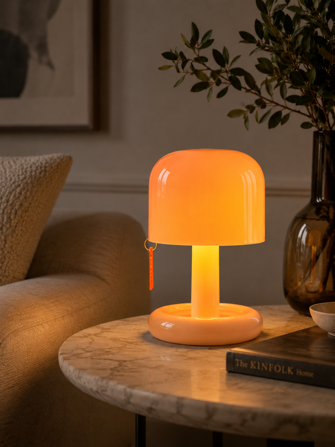 A glowing mushroom-shaped table lamp with a warm orange light, placed on a round marble side table in a cozy living room setting next to a sofa and a dark glass vase.