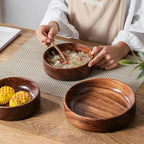 Person eating from a wooden bowl on a wooden table with other wooden bowls and food items.