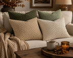 Four textured throw pillows in cream and sage green arranged on a light beige sofa, with a chunky knit cream throw blanket draped over the left side. A wooden tray with a mug and candle sits on a coffee table in the foreground, and framed wall art and a lamp are visible in the background.