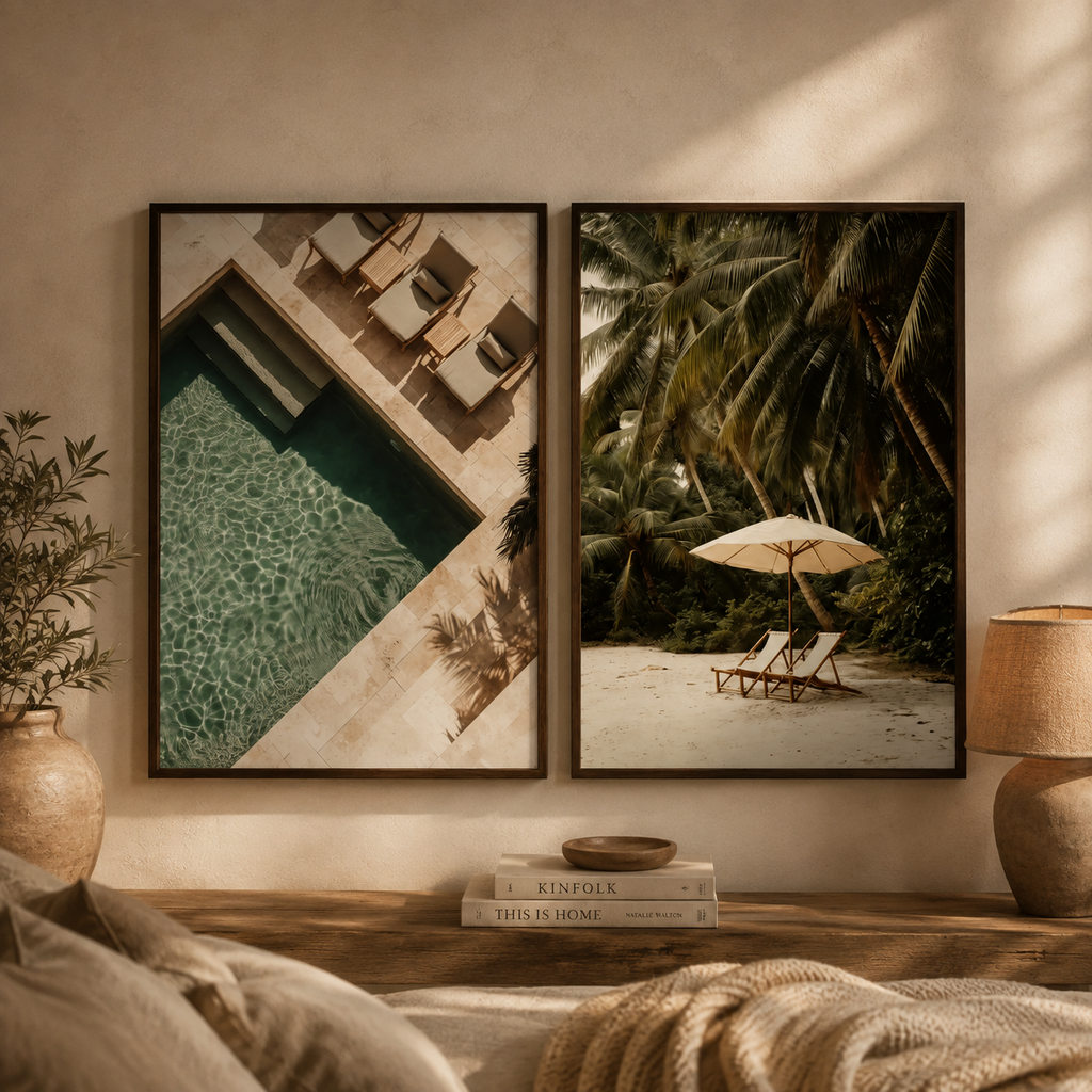 Two framed wall art prints hang on a beige wall above a wooden console. The left print shows an overhead poolside scene with lounge chairs, and the right print shows a tropical beach scene with palm trees, lounge chairs, and an umbrella.