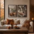 Two framed art prints with warm autumnal tones hang on a beige wall above a cream sofa styled with brown and beige pillows, a rust throw blanket, and a wooden coffee table in a cozy living room.