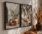 Two framed wall art prints hang on a sunlit beige wall. The left print shows terracotta vases with greenery, and the right print shows a table still life with shadows.