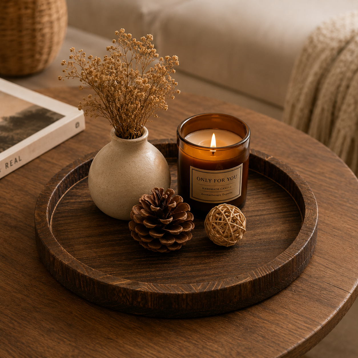 A lit amber glass jar candle with a cream label reading 'ONLY FOR YOU' sits on a round dark wood tray, styled with a small vase of dried flowers, a pinecone, and a woven ball on a wooden table in a cozy living room setting.