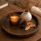 A round dark wood tray styled on a wooden table with a lit amber glass candle, a small speckled ceramic vase holding dried stems, a pinecone, and dried leaves.