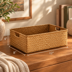 A woven rectangular storage basket with cutout handles sits on a wooden table in a styled room setting. White dimension lines and text show the basket's measurements: 12 inches wide, 8 inches deep, and 4 inches tall.