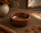 A round wooden bowl with a rich dark brown grain pattern, set on a rustic wooden table with autumn leaves and a ceramic vase in the background.