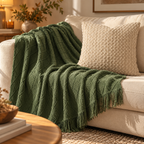 A textured olive green throw blanket with fringe is draped over a cream-colored sofa next to a woven beige pillow in a warm, sunlit living room setting.