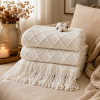 Three folded cream-colored textured throw blankets with fringe are stacked on a beige sofa, styled with dried flowers in a vase, a lit candle, and a cotton boll.